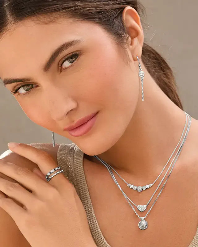 A close-up lifestyle shot of a woman wearing a silver jewelry set, including a layered necklace with heart pendants, a drop earring, and stacked rings.