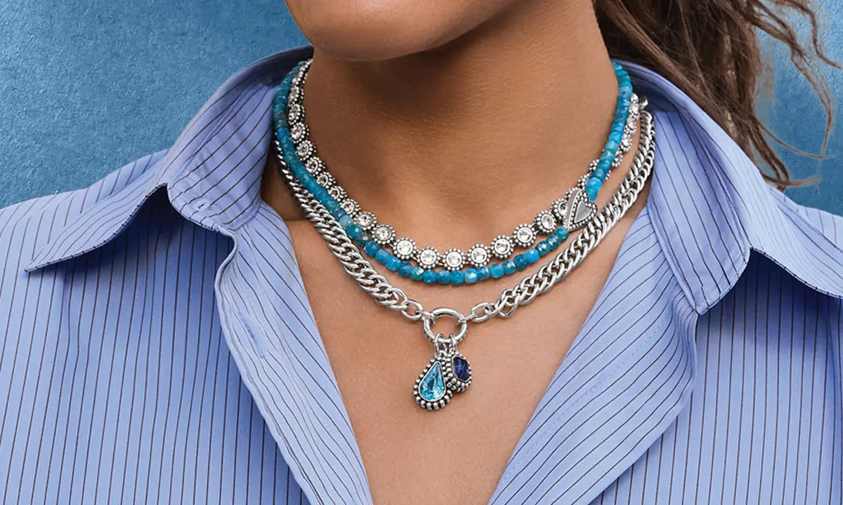 A close-up lifestyle shot of a woman wearing a layered jewelry set featuring a silver chain necklace with turquoise and crystal accents, a matching beaded necklace, and a silver drop earring.