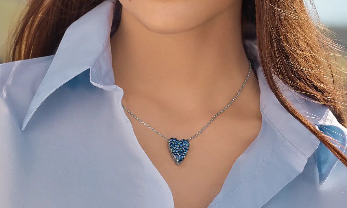 A close-up of a woman wearing a silver chain necklace with a heart-shaped pendant covered in sparkling blue stones, paired with a light blue collared shirt.