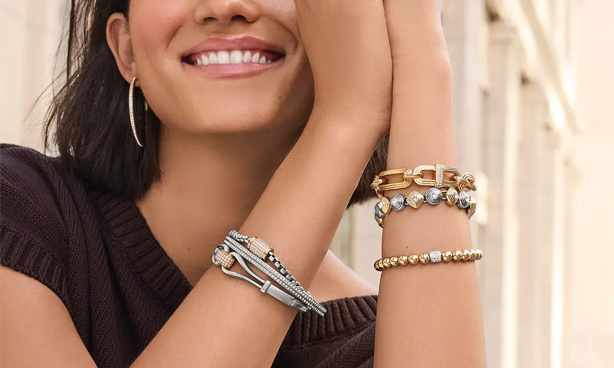 A smiling woman wearing multiple gold and silver bracelets. There are bangles with crystals, beaded stretch bracelets, and chain bracelets.