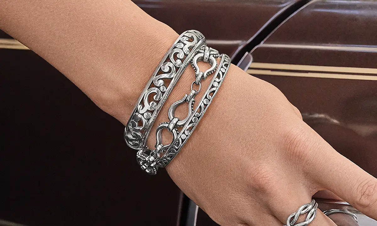 A model's wrist wearing three silver scroll bracelets in front of a brown car.