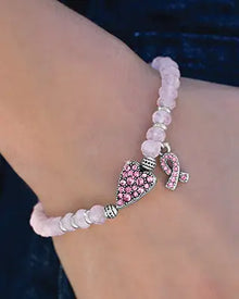 A close up shot of a pink beaded bracelet with a heart with pink crystals and a breast cancer charm with pink crystals. 
