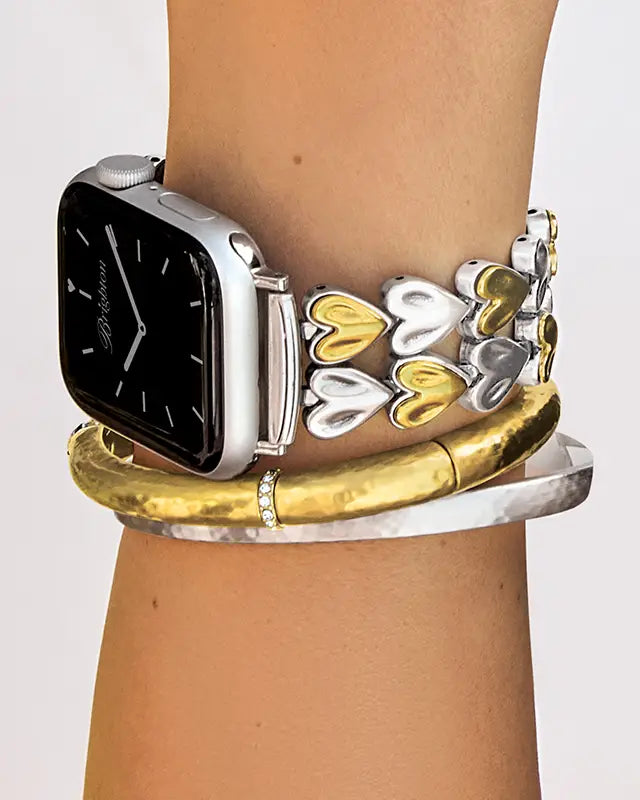 A close-up of a person's wrist wearing a silver smartwatch with a decorative bracelet featuring interlocking silver and gold hearts, layered over a thick gold bangle and a silver bangle.