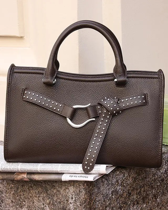 A dark brown pebbled leather handbag with double top handles, a front strap detail with silver studs, and a large silver metal ring, resting on a newspaper against a neutral background.