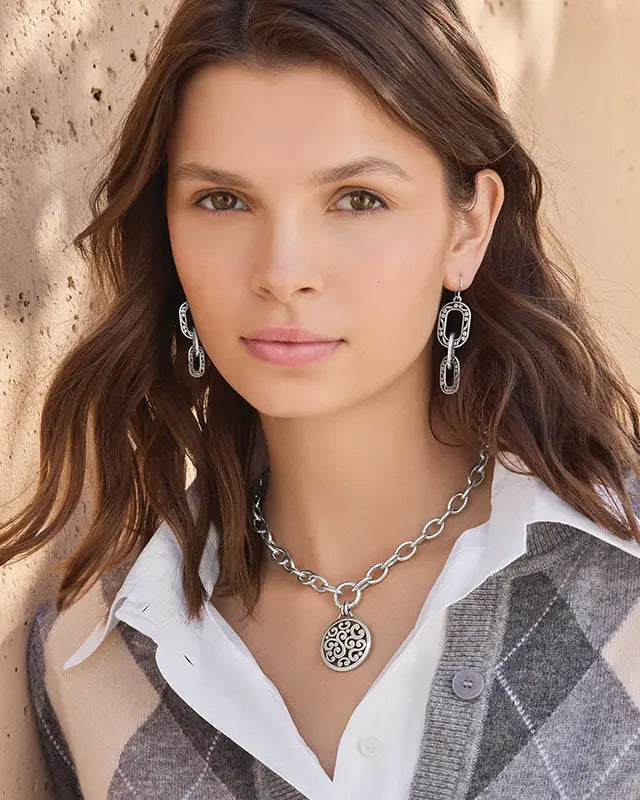 A close-up portrait of a woman wearing a silver chain necklace with a circular filigree pendant, matching silver drop earrings, and a gray plaid cardigan over a white collared shirt.
