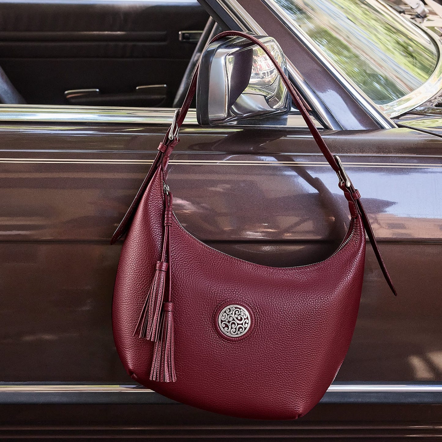 Brighton burgundy leather hobo handbag with silver-tone medallion and leather tassels, hanging on a vintage car door