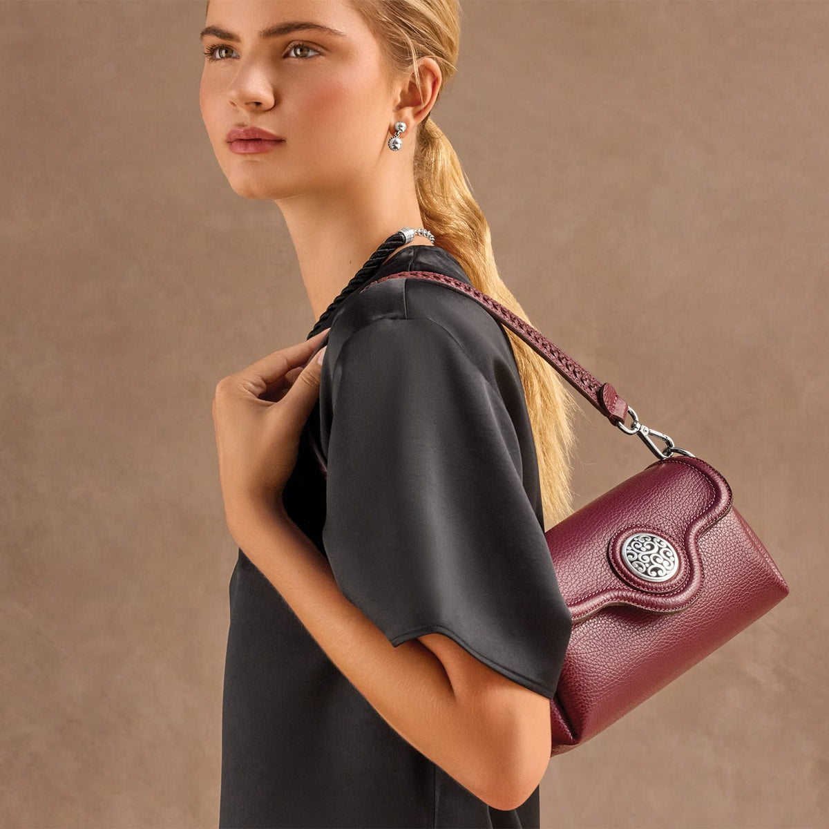 Model with a Brighton burgundy leather shoulder handbag featuring a silver-tone medallion accent and braided strap