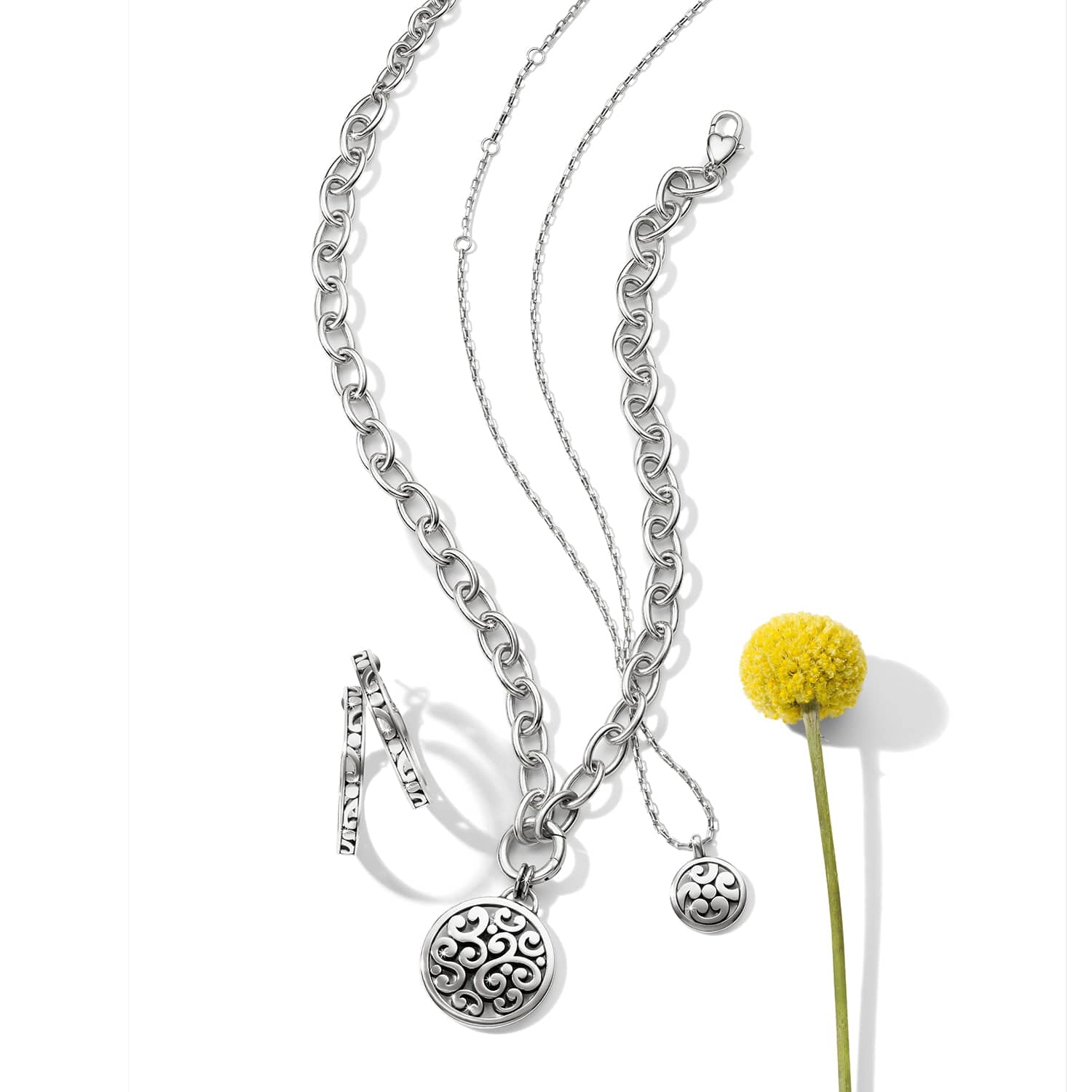 Brighton silver-plated jewelry set styled with a yellow craspedia flower, featuring chain necklaces, scroll pendants, and hoop earrings