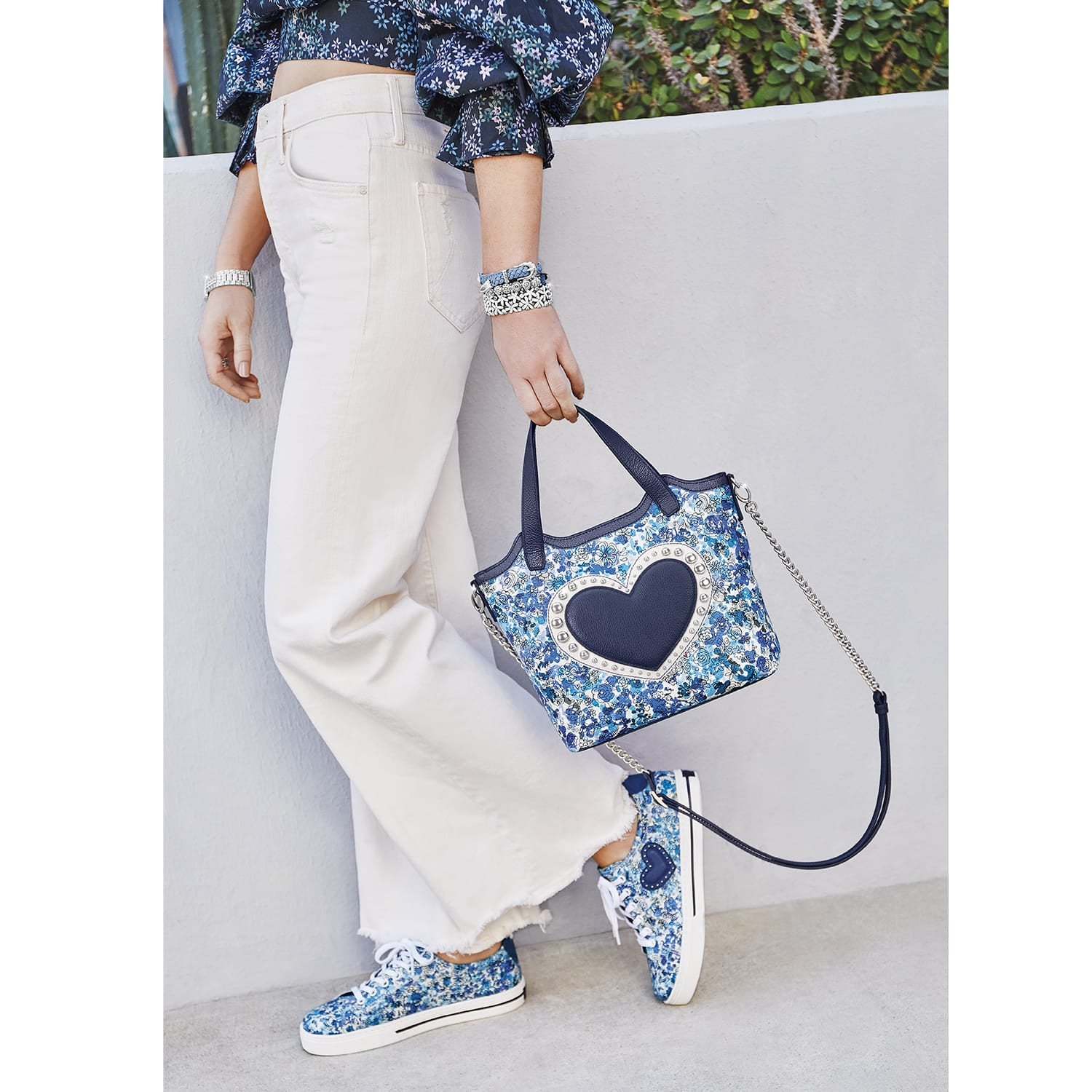 Model with a Brighton blue floral leather handbag featuring navy heart medallion, studded trim, and chain strap