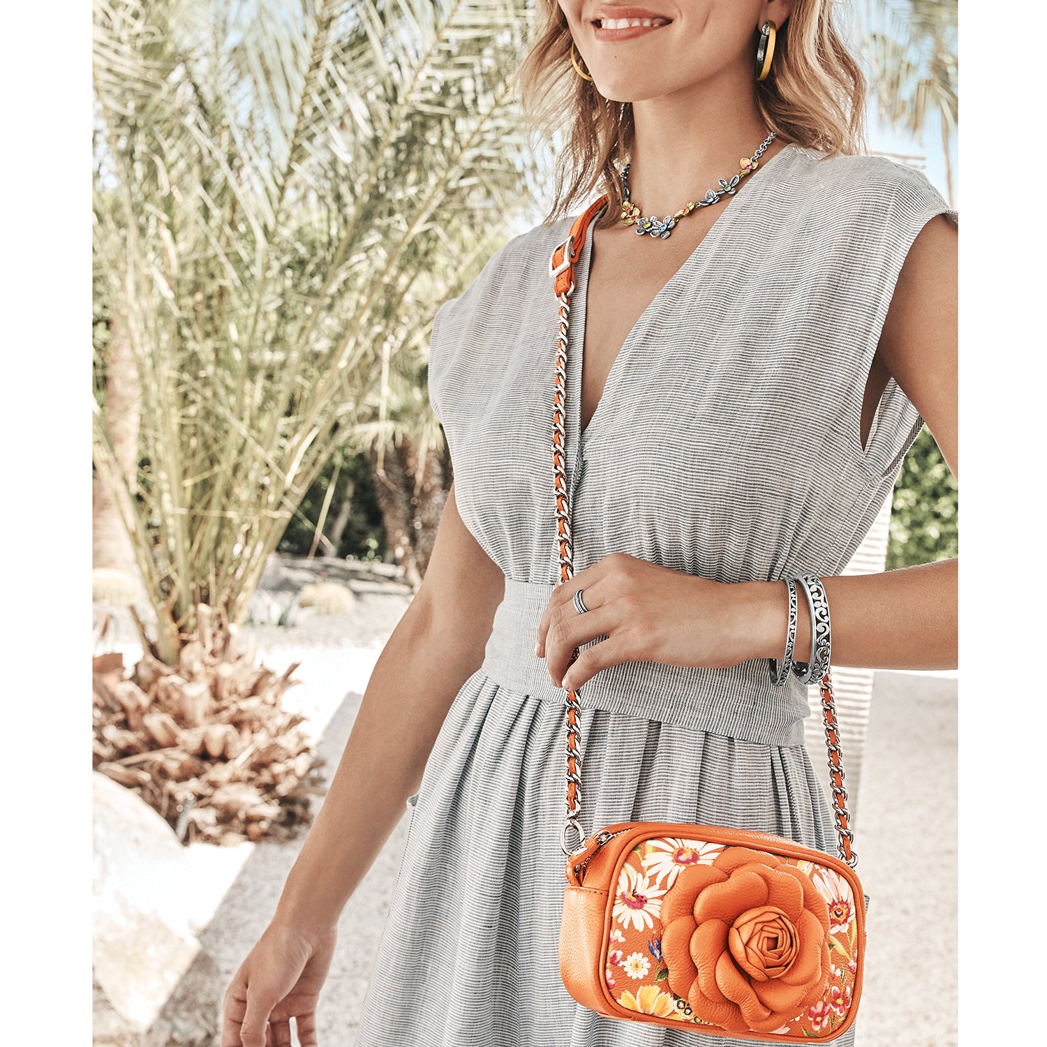 Model with a Brighton orange crossbody handbag featuring a floral print and dimensional rose with silver-tone chain strap
