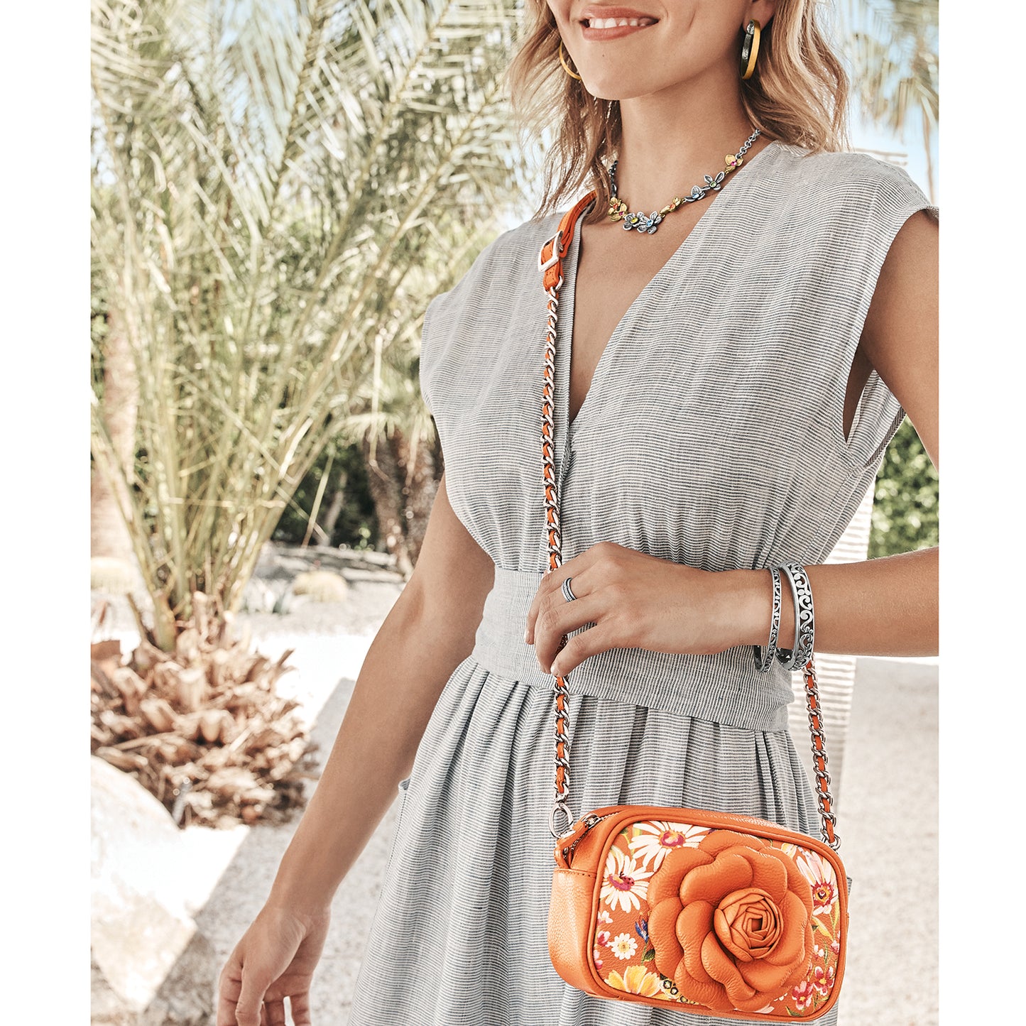 Model with a Brighton orange crossbody handbag featuring a floral print and dimensional rose with silver-tone chain strap