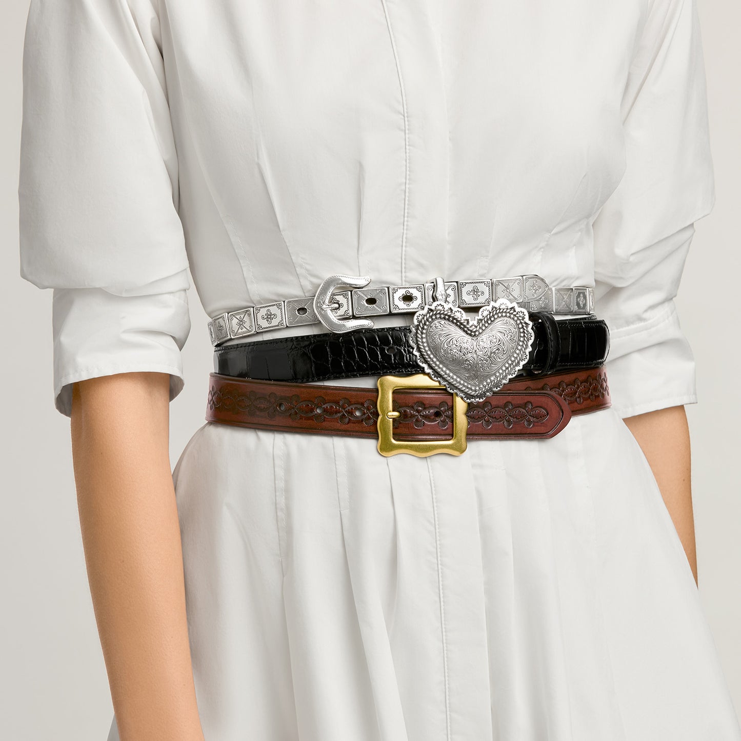 Model wearing three stacked Brighton leather belts with silver studded strap, black croc-embossed heart buckle, and brown tooled design
