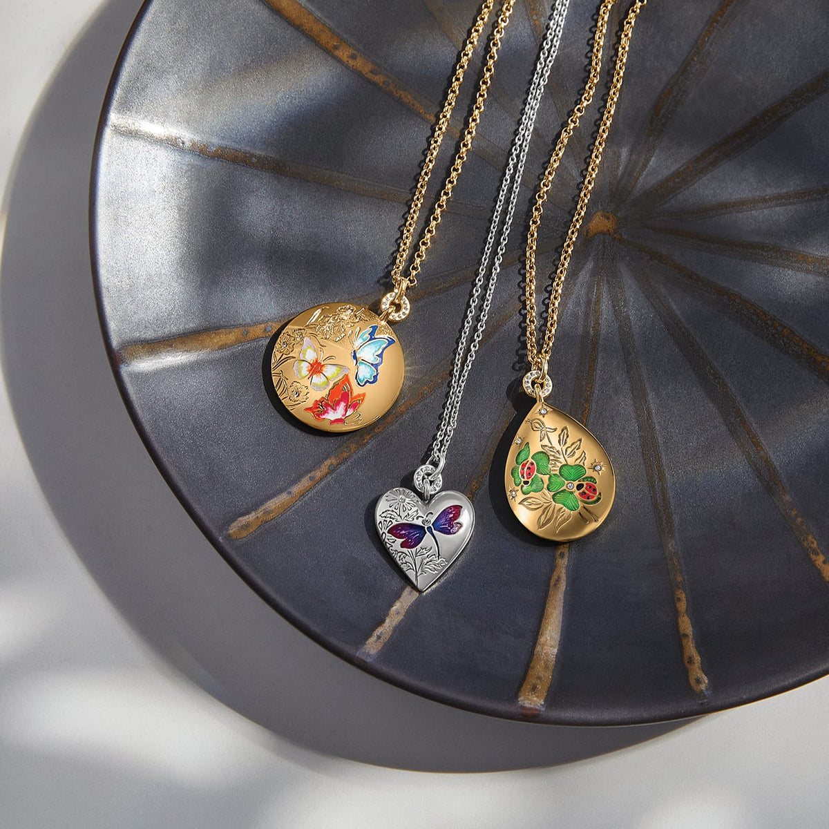 Brighton locket necklaces styled with enamel butterfly, ladybug, and dragonfly motifs on a metallic round tray