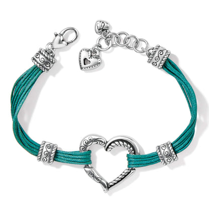 Brighton bracelet with teal cords, silver-plated heart centerpiece, scrolled beads, and heart charm on a white background