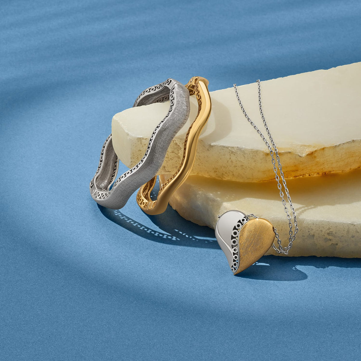 Brighton two-tone bangles and heart pendant necklace styled with stone slabs on a blue background