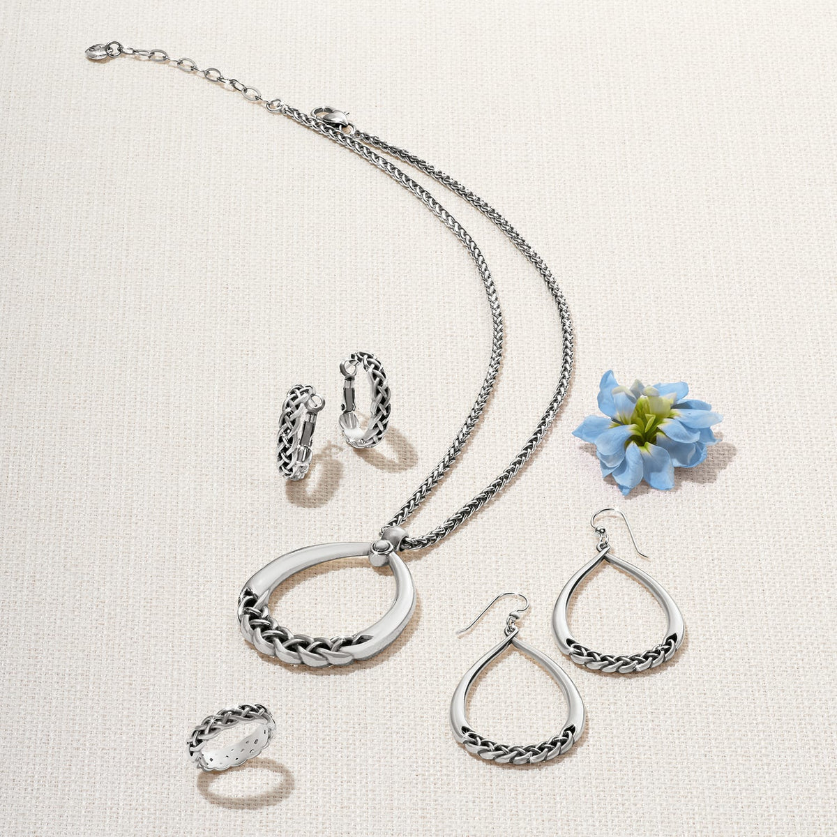 Brighton silver-plated jewelry set styled with a blue flower, featuring braided pendant necklace, teardrop earrings, hoops, and ring