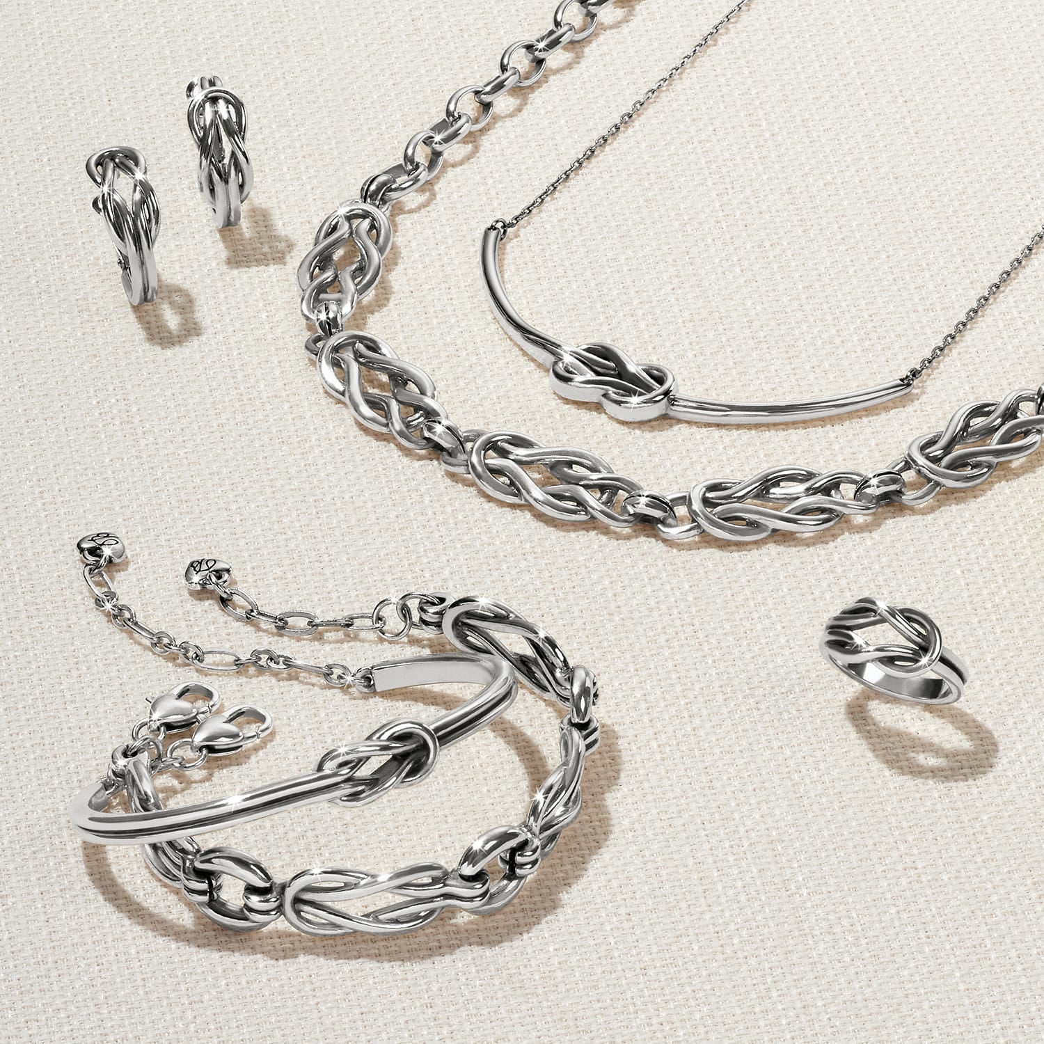 Brighton silver-plated jewelry set styled with knotted bracelet, necklace, earrings, and ring on a neutral woven background