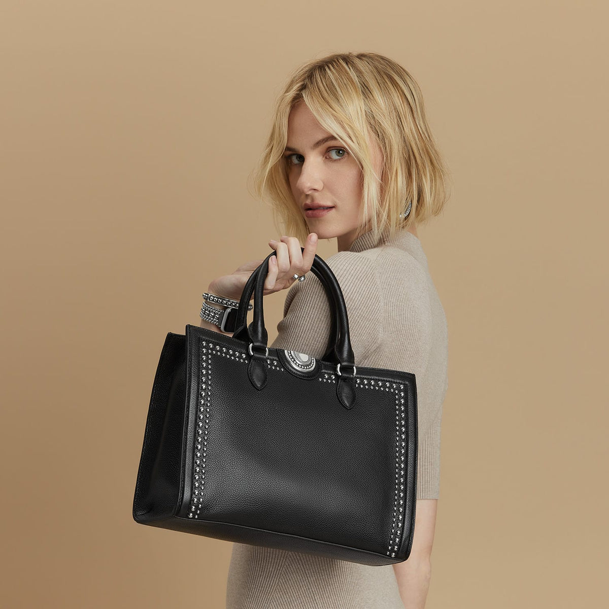 Model with a Brighton black leather handbag featuring silver-tone studded trim and a circular medallion accent