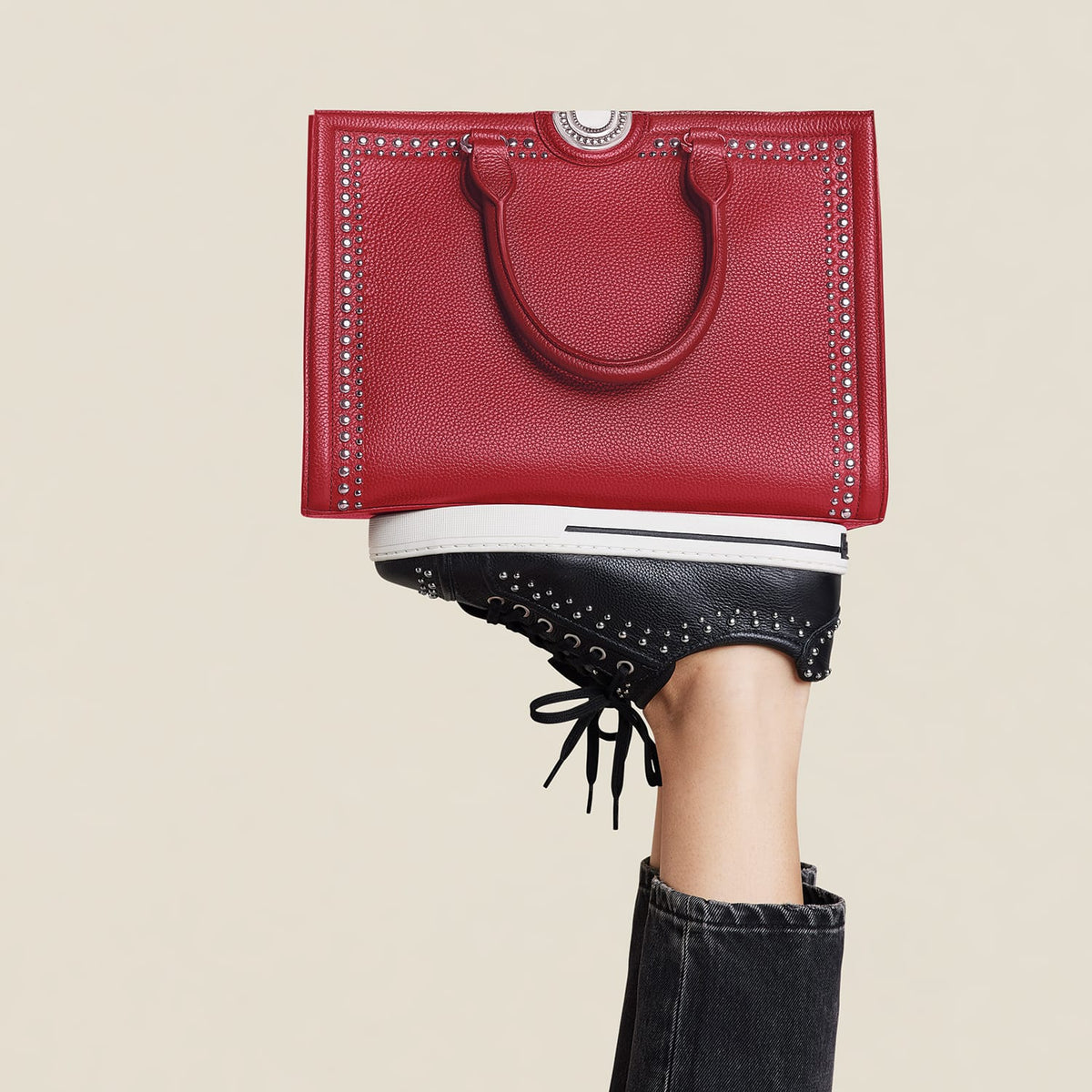 Brighton red studded leather tote handbag balanced on model's foot wearing black studded lace-up sneakers
