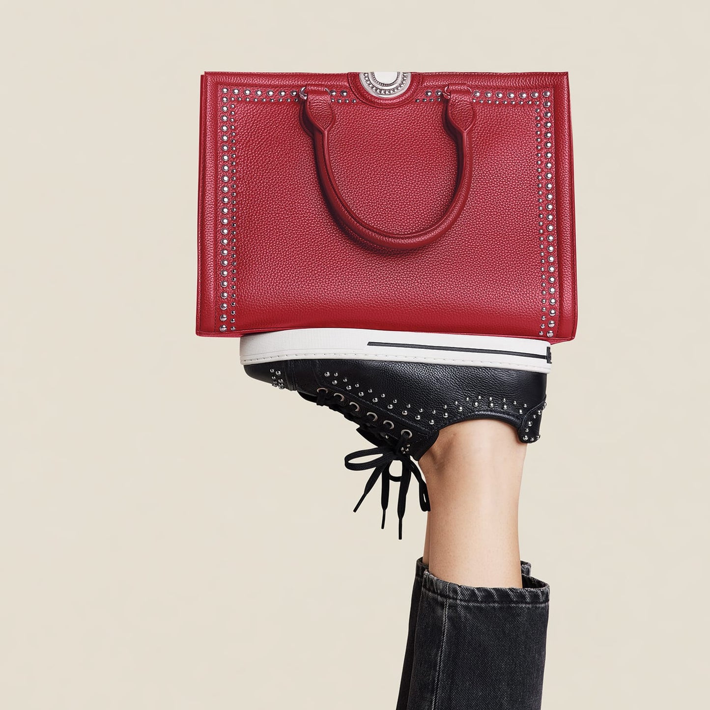 Brighton red studded leather tote handbag balanced on model's foot wearing black studded lace-up sneakers