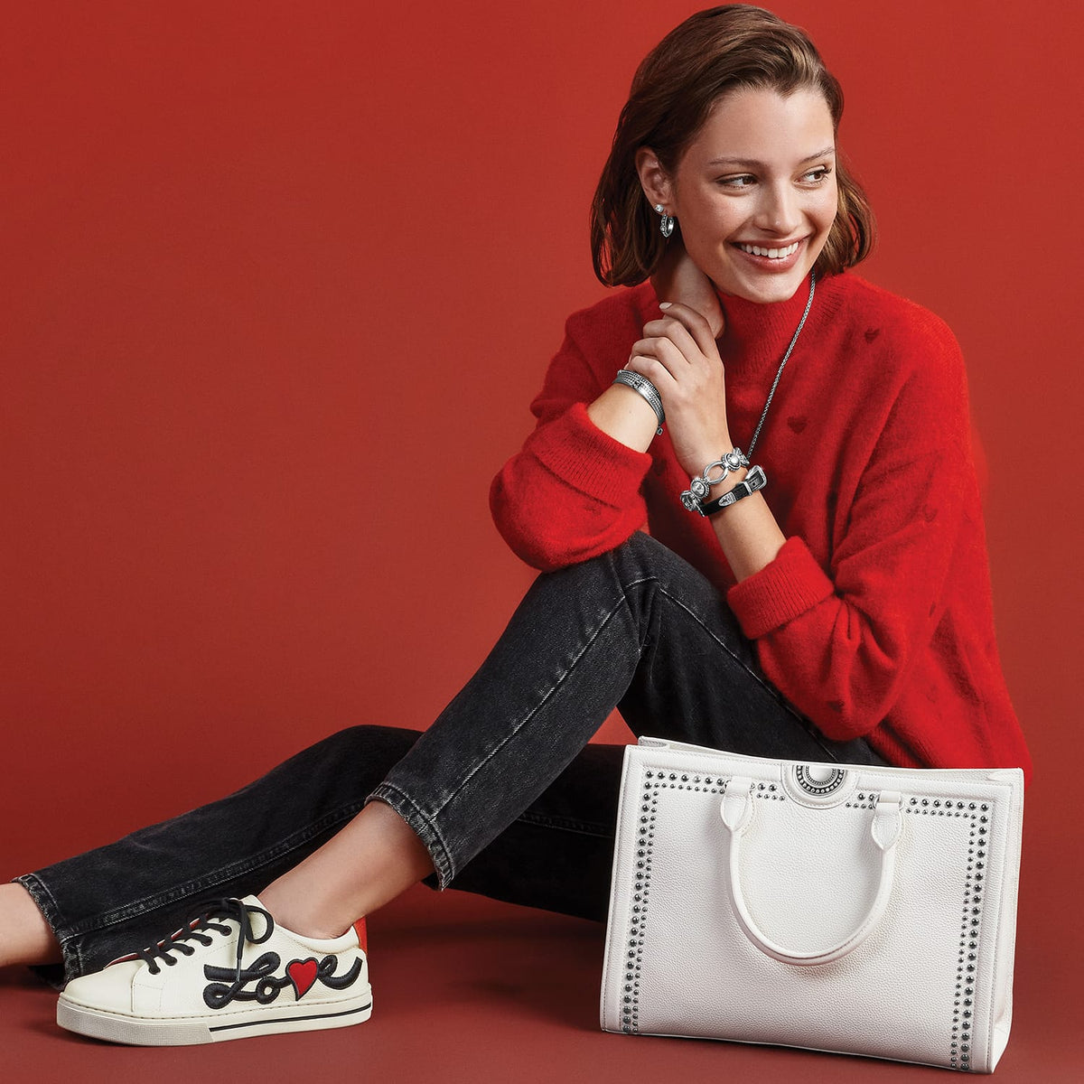 Model with a Brighton white studded leather tote handbag, styled with coordinating Brighton accessories and love-print sneakers