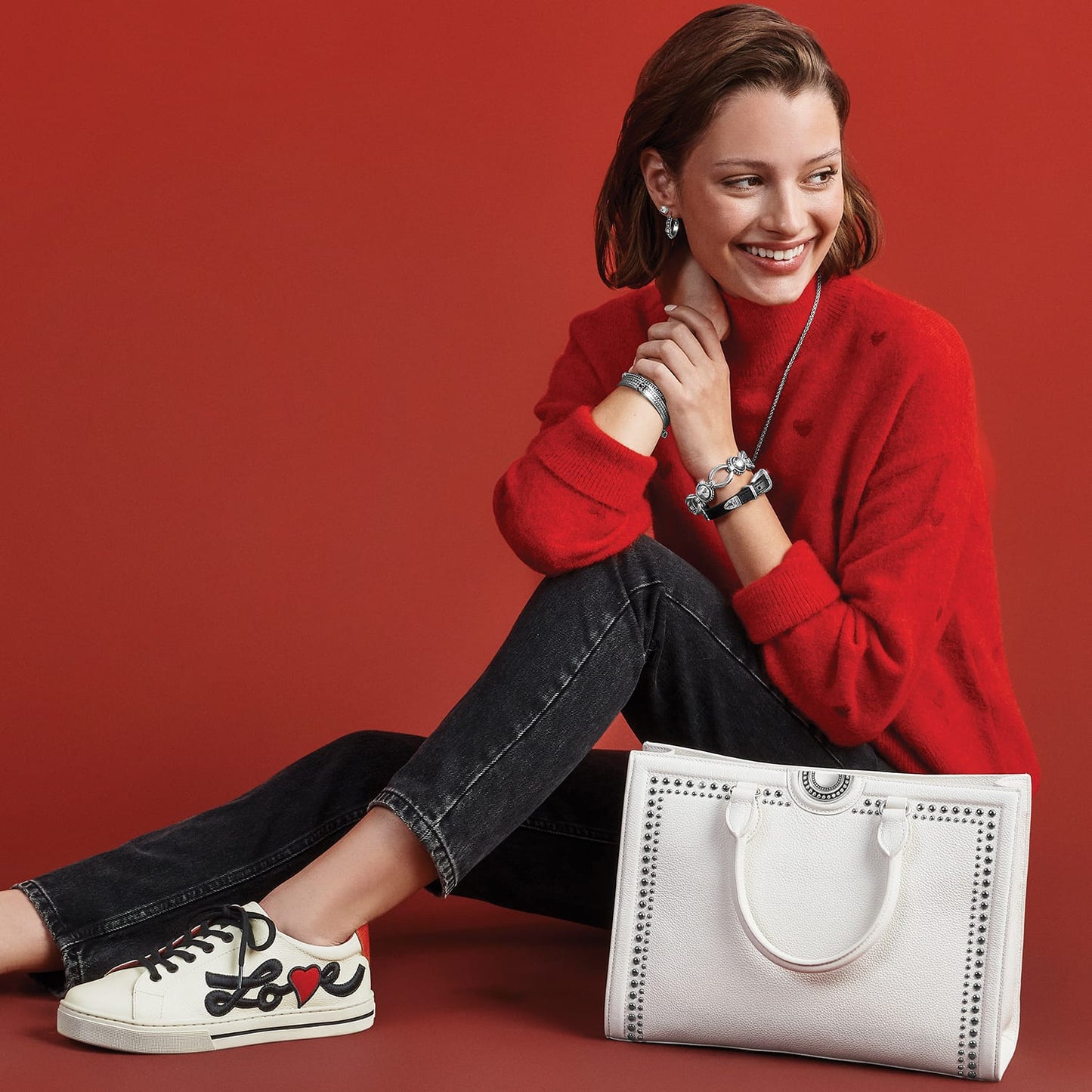 Model with a Brighton white studded leather tote handbag, styled with coordinating Brighton accessories and love-print sneakers
