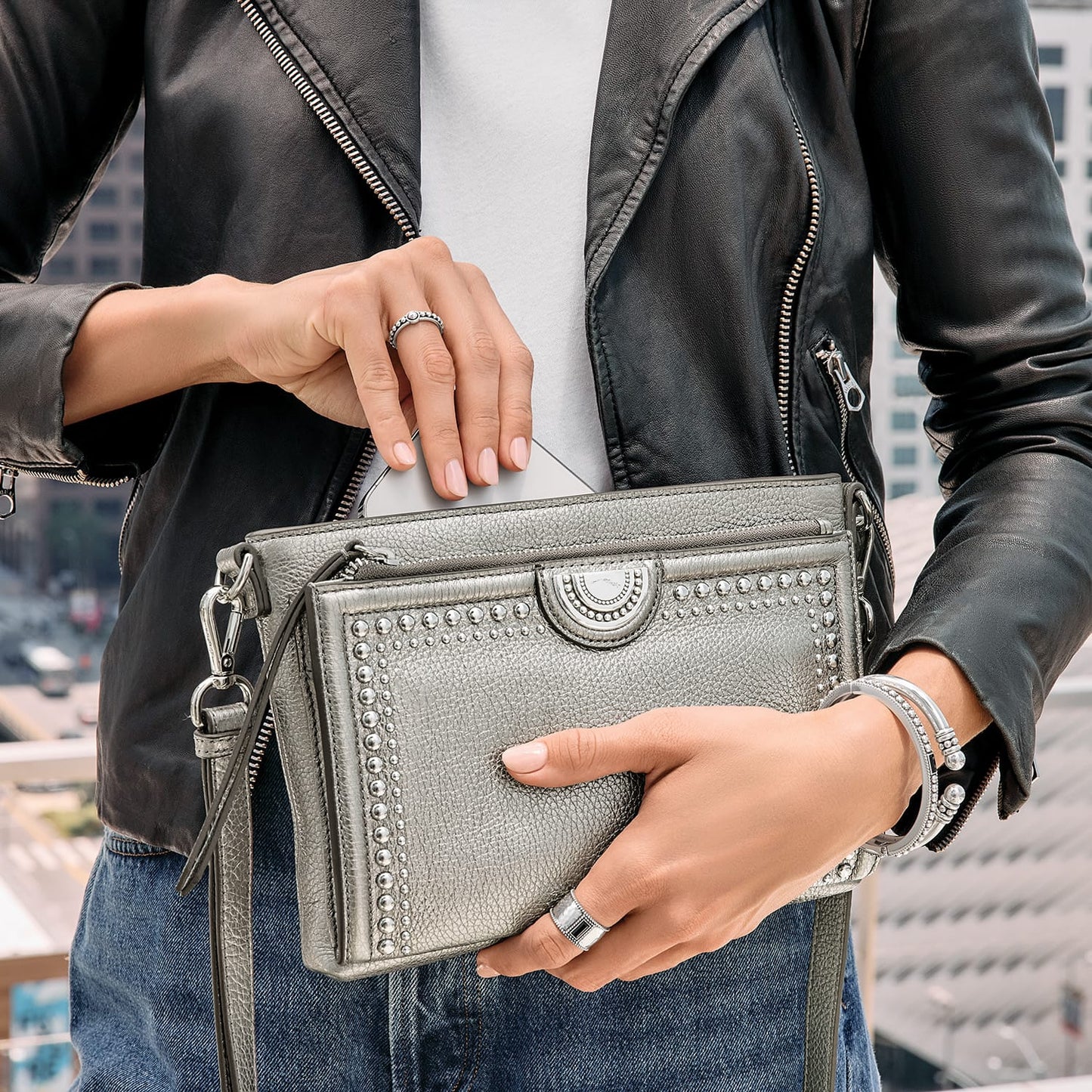 Model with a Brighton metallic silver studded handbag placing a phone inside, styled with silver bangles