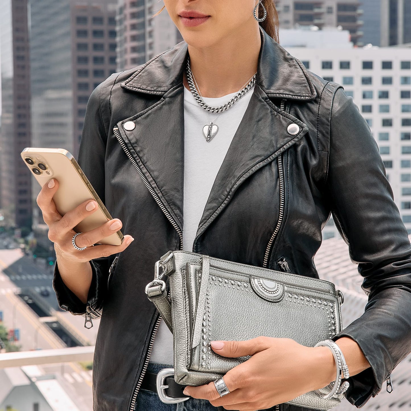Model with a Brighton metallic silver crossbody handbag with studded trim and medallion accent, holding a phone