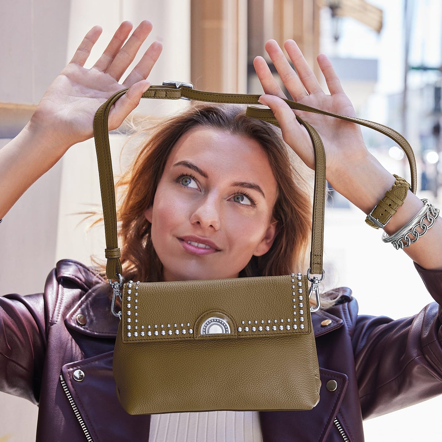 Model holding a Brighton olive green leather handbag with silver-tone studded trim and adjustable crossbody strap