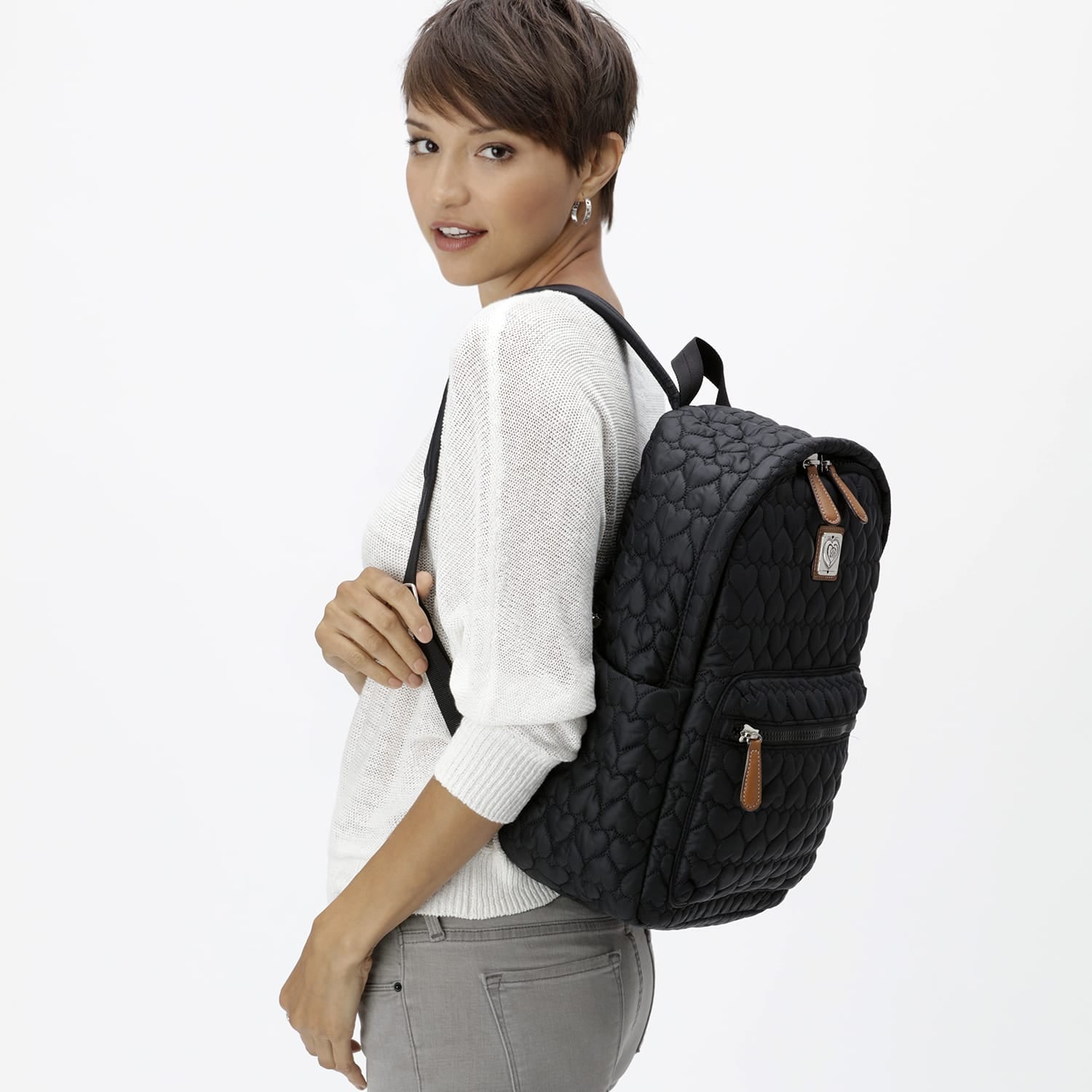 Model with a Brighton black quilted backpack featuring heart-patterned stitching, tan leather zipper pulls, and silver-tone hardware