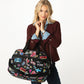 Model with a Brighton black duffle handbag featuring colorful graphic print of hearts, stars, and travel motifs