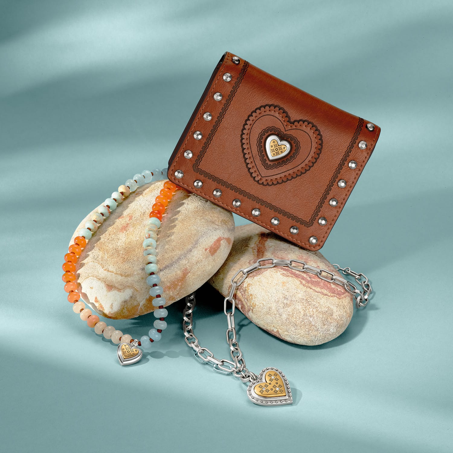 Brighton heart wallet styled with beaded and silver-tone chain bracelets on stones against a teal background