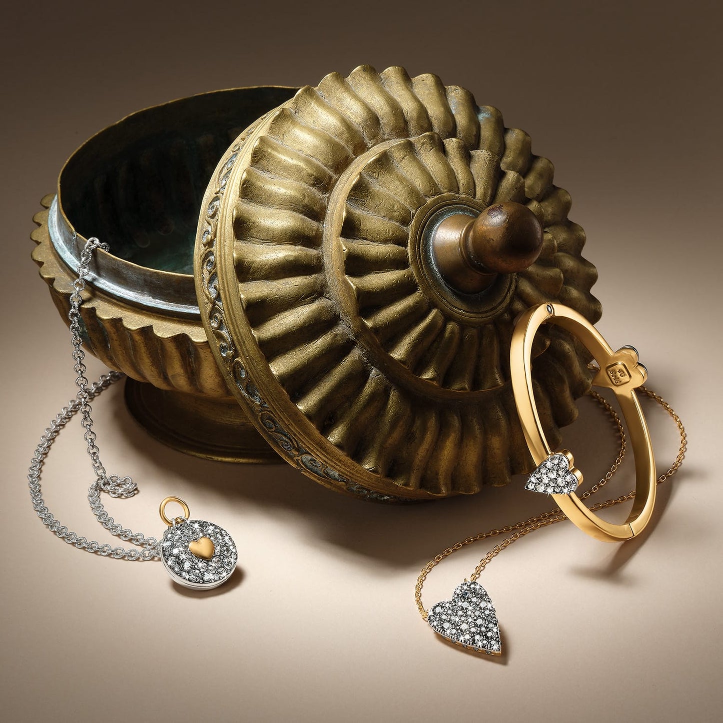 Brighton two-tone crystal heart necklaces and bangle styled with an ornate brass vessel on a neutral background