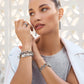 A woman gazes away, showcasing the Pretty Tough Droplet Hinged Bangle with matching silver jewelry, against a white, patterned background. She wears rings, bracelets, and dangling earrings, paired with a white jacket and gray top.