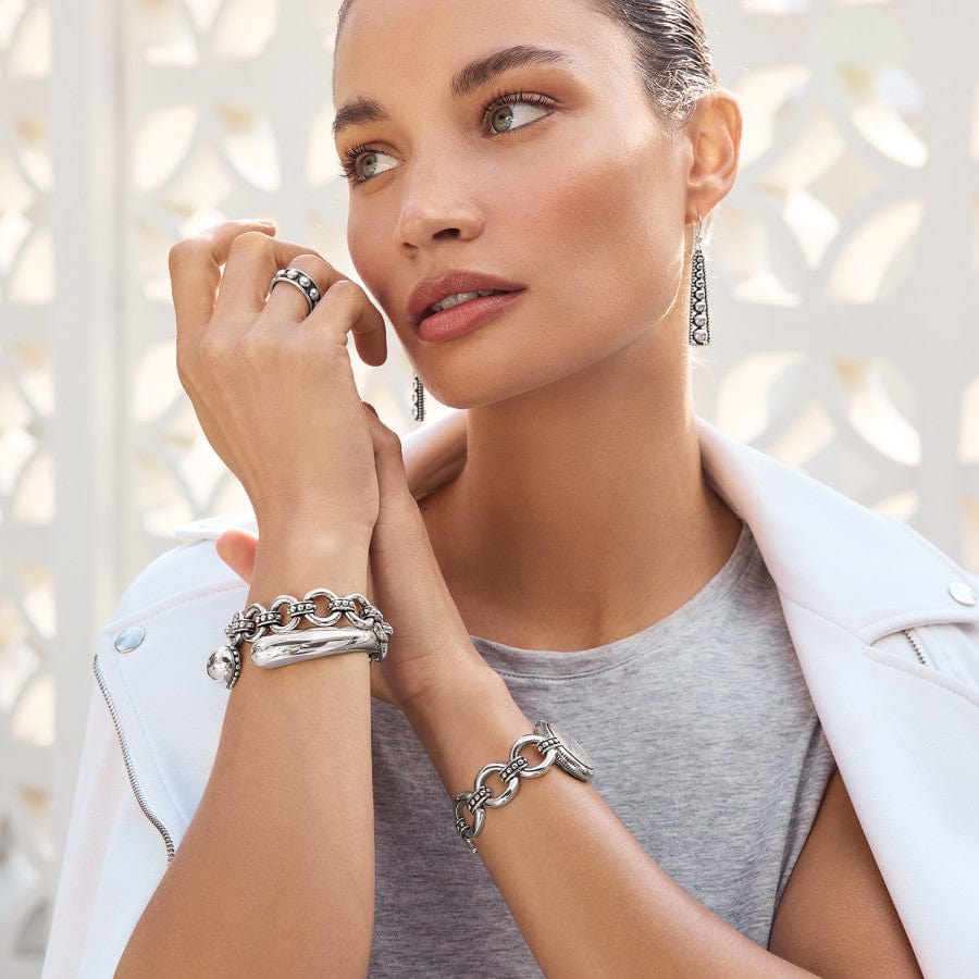A woman gazes away, showcasing the Pretty Tough Droplet Hinged Bangle with matching silver jewelry, against a white, patterned background. She wears rings, bracelets, and dangling earrings, paired with a white jacket and gray top.