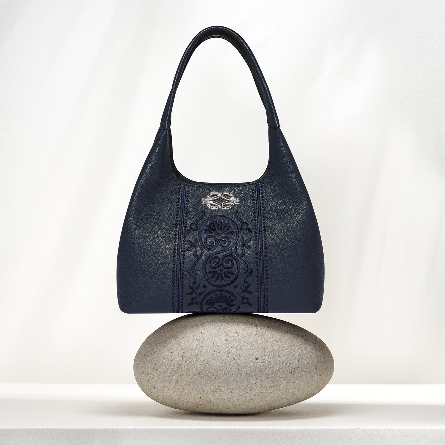 Brighton navy leather handbag with tonal scroll embroidery and silver-tone medallion, balanced on a stone on a white background