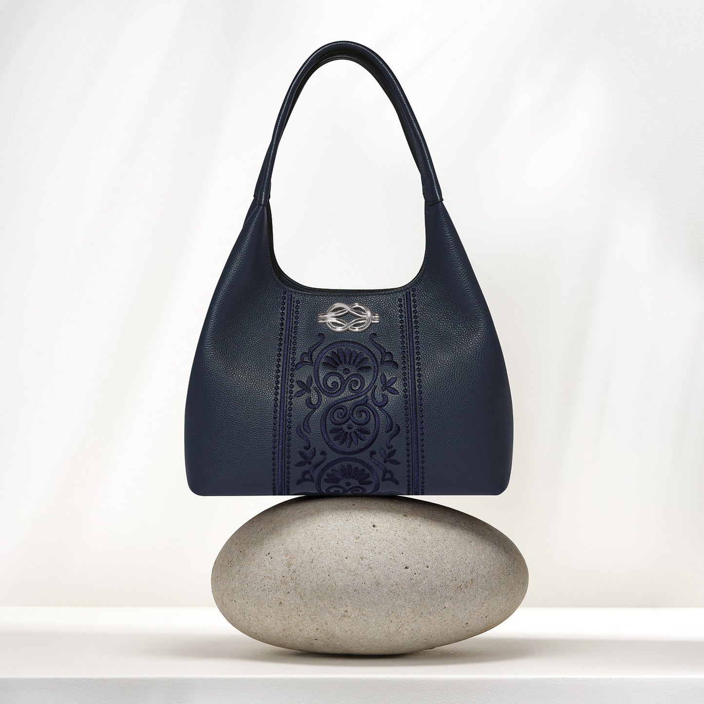 Brighton navy leather handbag with tonal scroll embroidery and silver-tone medallion, balanced on a stone on a white background