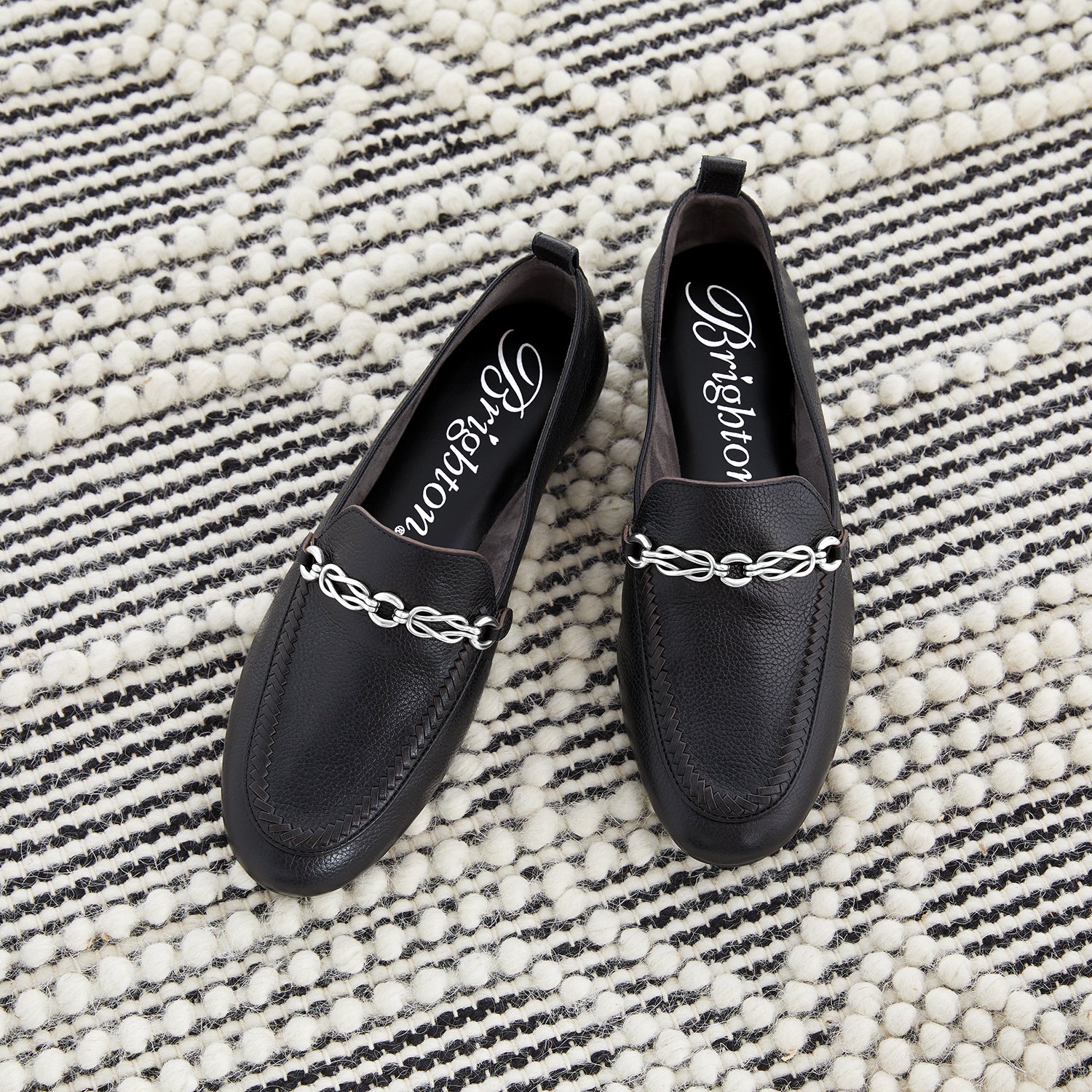 Brighton black leather loafers styled with silver-tone chain hardware and woven detail on a striped woven rug