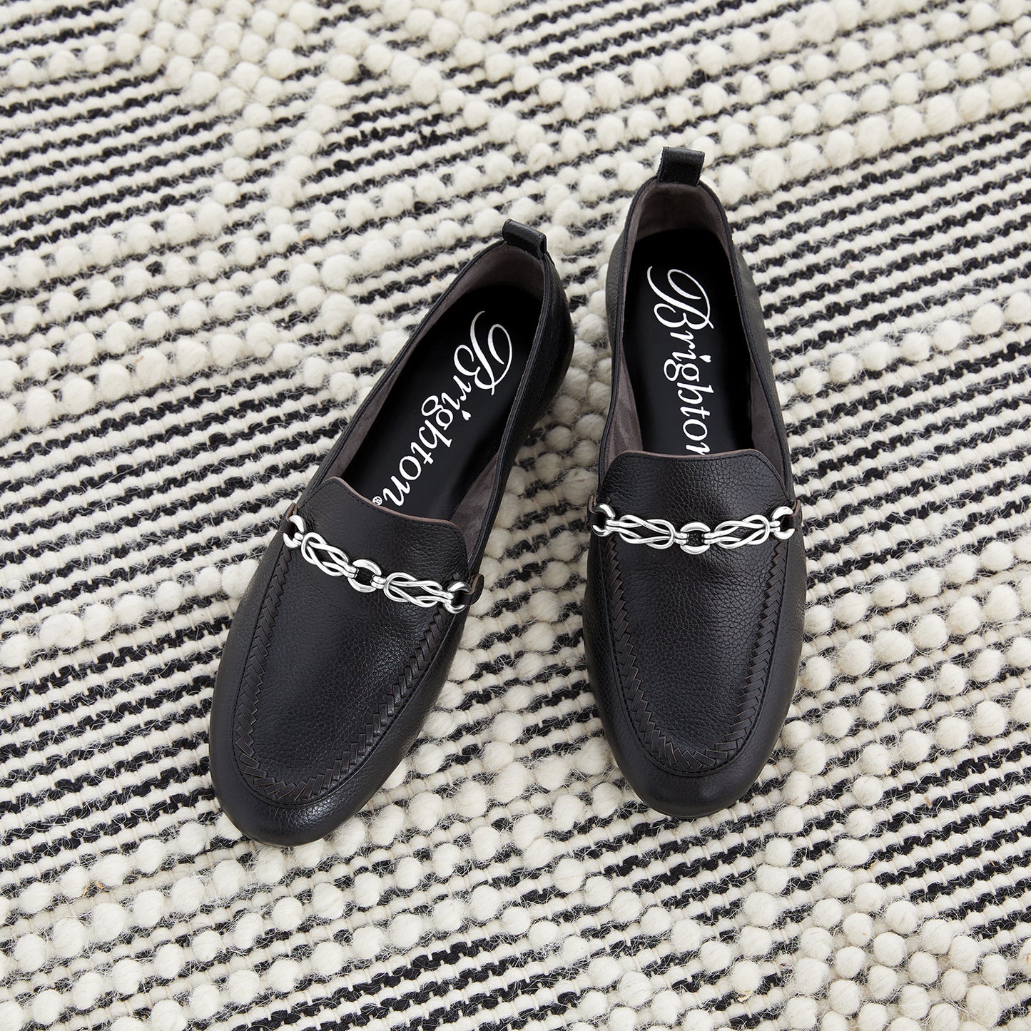 Brighton black leather loafers styled with silver-tone chain hardware and woven detail on a striped woven rug