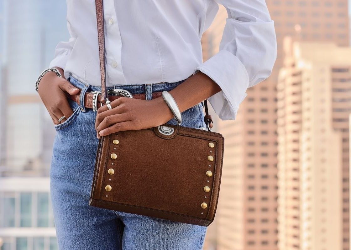 A person wearing a white button-up shirt and blue jeans carries a brown leather crossbody bag with gold-tone studs and a silver bangle, set against a blurred cityscape background.