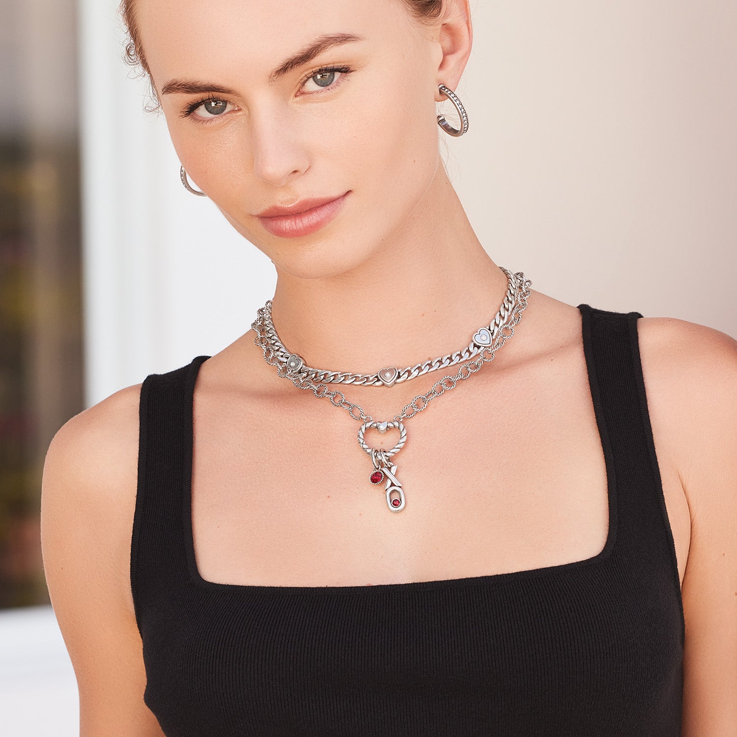 Model wearing layered Brighton silver-plated chain necklaces with heart accents and red crystal charms, paired with hoop earrings