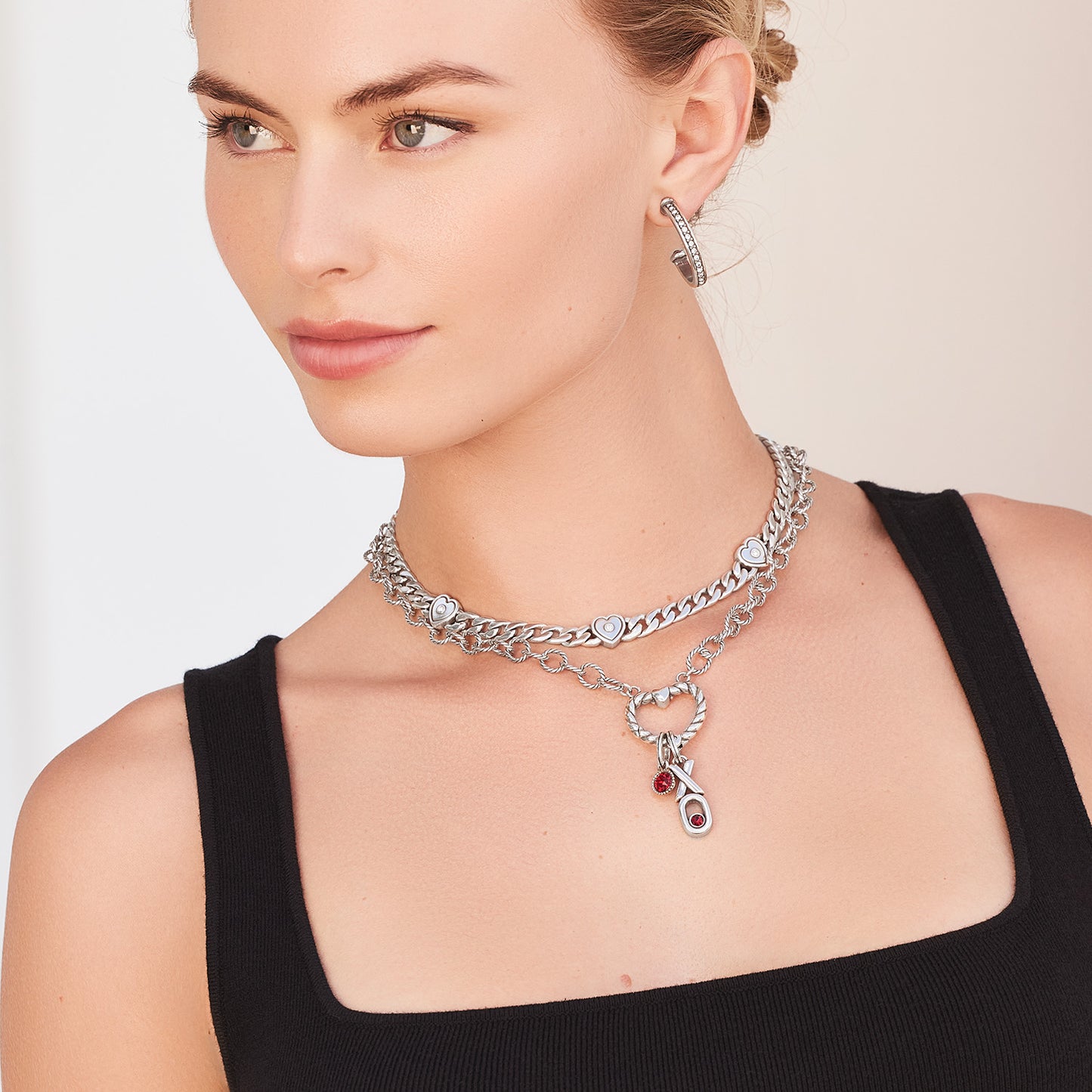 Model wearing layered Brighton silver-plated chain necklaces with heart stations, red crystal charm, and coordinating Brighton accessories