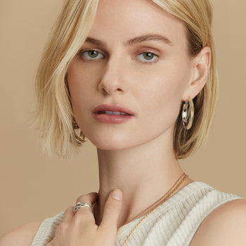 A model wearing the Interlok Heirloom Hoop Earrings with short, wavy brown hair is smiling slightly and a pink top against a white background.