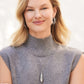 A smiling model wearing the Pretty Tough Droplet Necklace with silver hoop earrings, dressed in a grey sleeveless top with a turtleneck, in a softly lit neutral setting.
